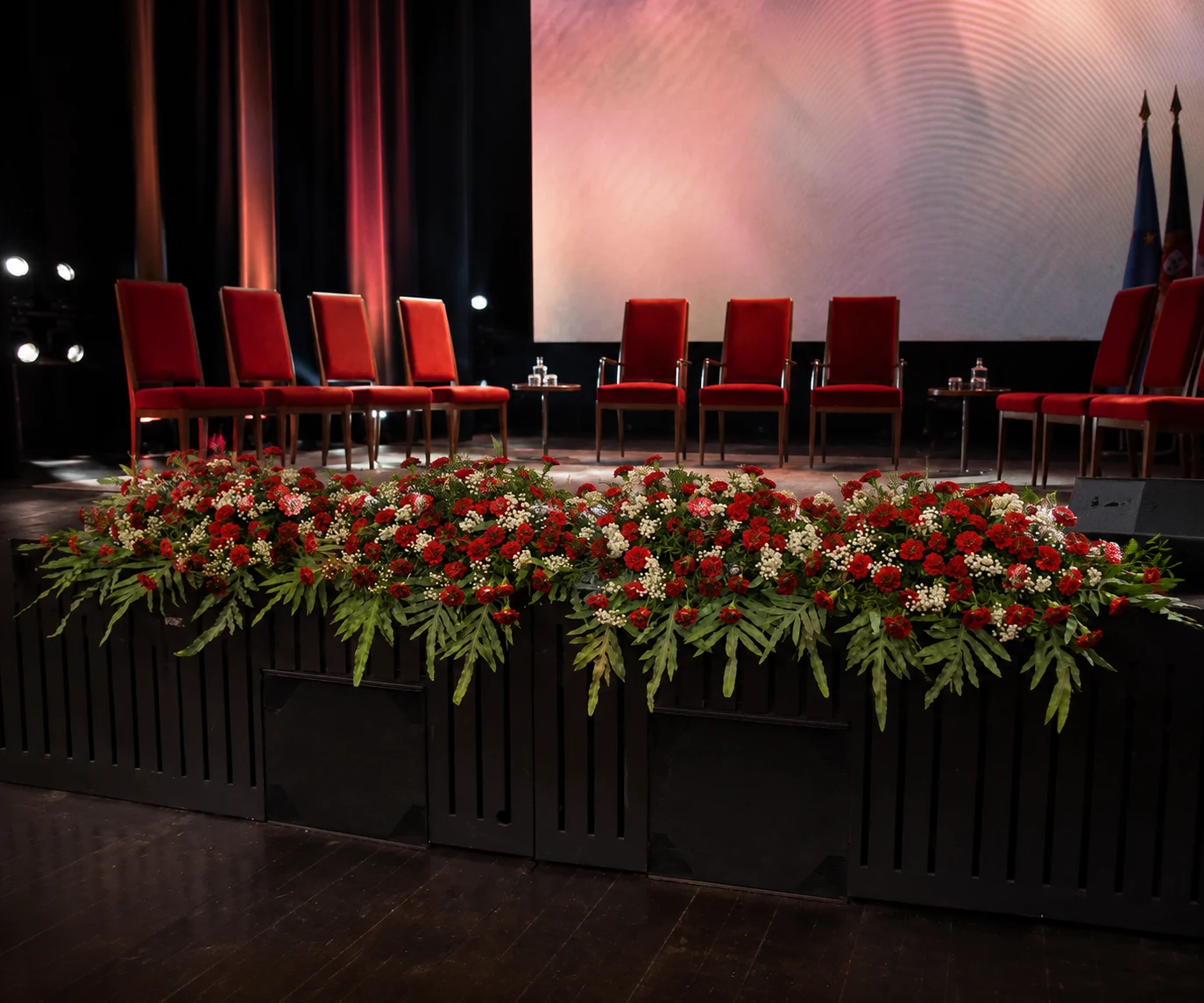 Decoração floral para evento com arranjo vermelho na frente de um palco preparado para cerimónia.